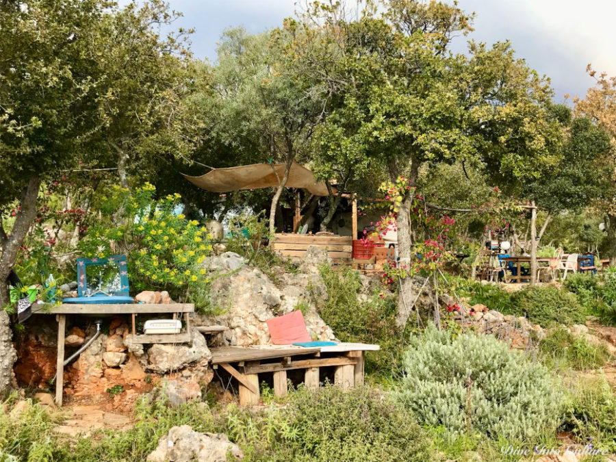 Paulas Permakultur-Paradies: kleine Waschstelle und Blick auf die Außenküche und den Essbereich zwischen bunt-blühenden Blumen und grünen Baumkronen, einbettet in die natürlichen Felsformationen